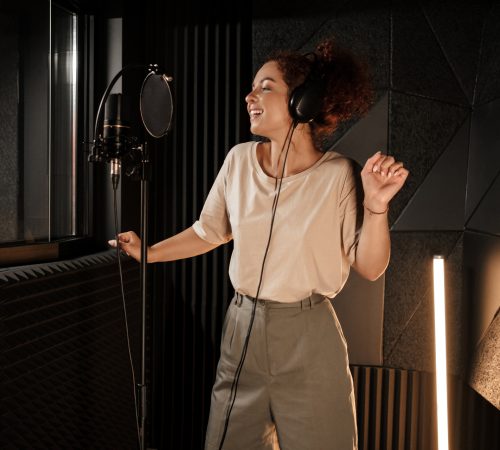 Charming female musician in headphones dreamily dancing while recording song in modern studio